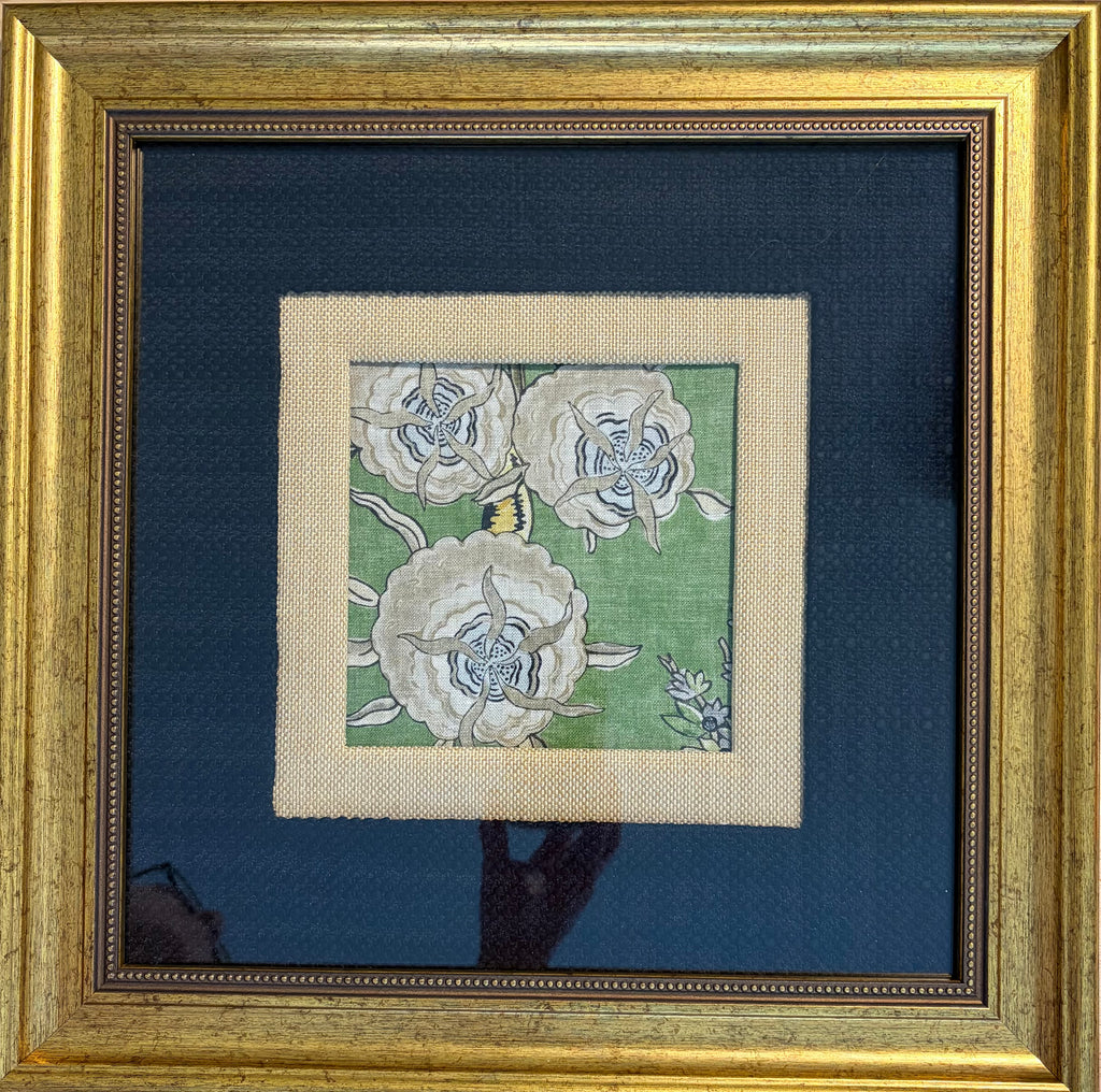 A framed floral needlepoint or embroidery piece featuring three large white flowers on a green background, surrounded by a beige mat and a wide dark blue mat, all enclosed in a gold frame.
