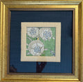 A framed floral needlepoint or embroidery piece featuring three large white flowers on a green background, surrounded by a beige mat and a wide dark blue mat, all enclosed in a gold frame.