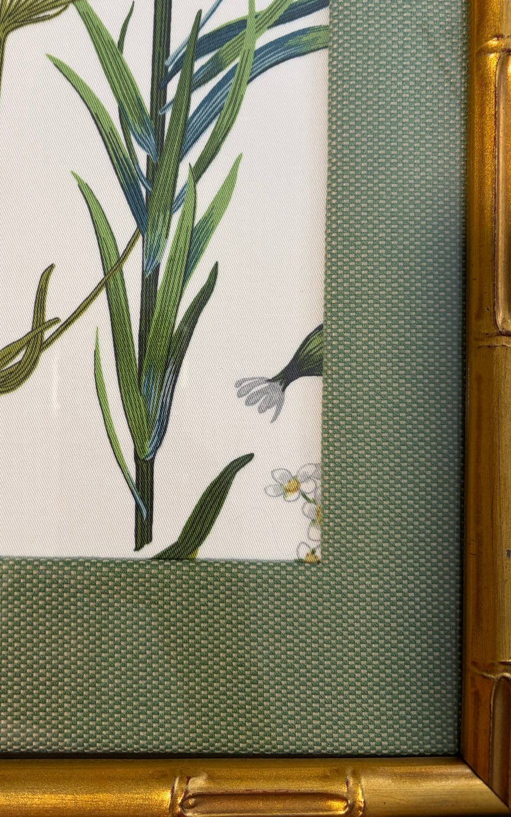 Lower-right detail of tall green leaves and small pale flowers beside the green mat and gold bamboo-style frame.