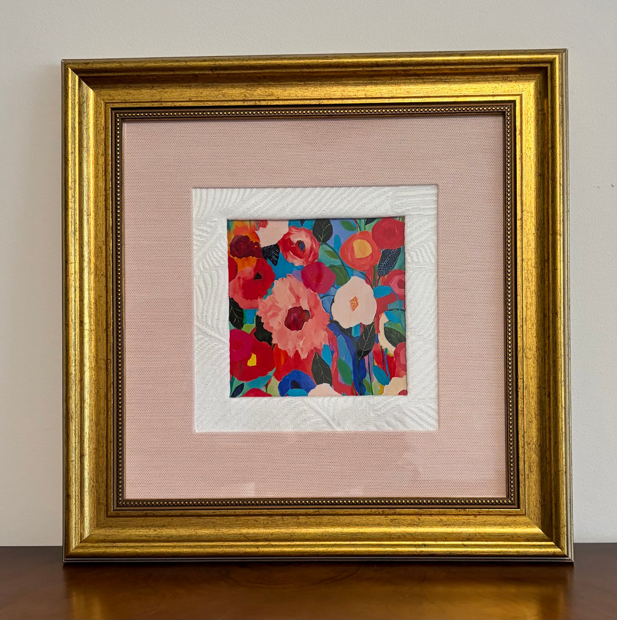 Framed floral wall art with a colorful abstract flower print in the center, surrounded by a white textured mat and a wide blush pink mat, all enclosed in a wide ornate gold frame.